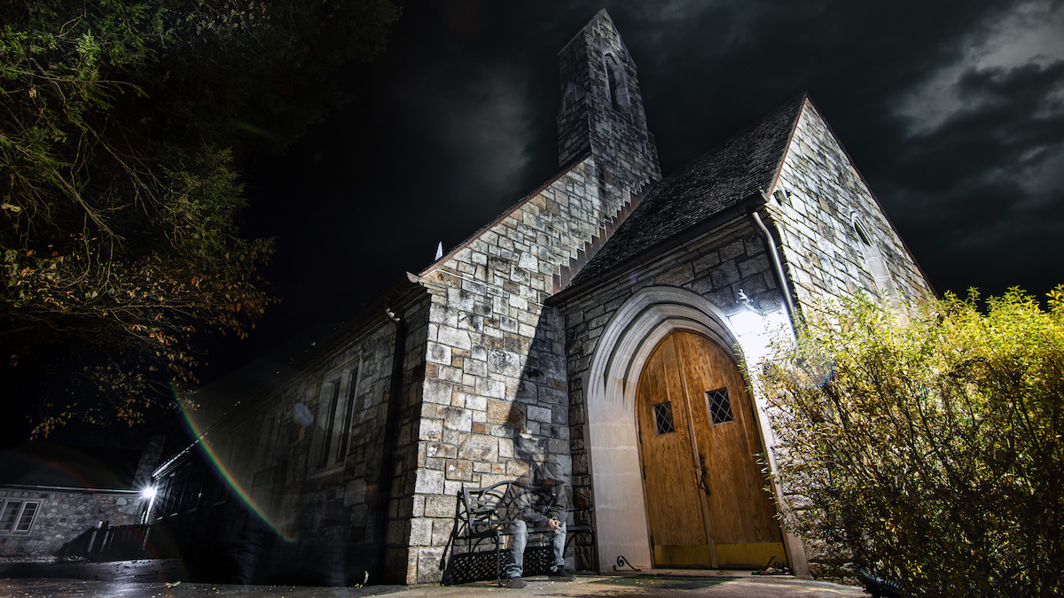 Night photo of beautiful dark sky with illuminated clouds. A historic stone First United Methodist Church in foreground with brown wooden door and foliage.