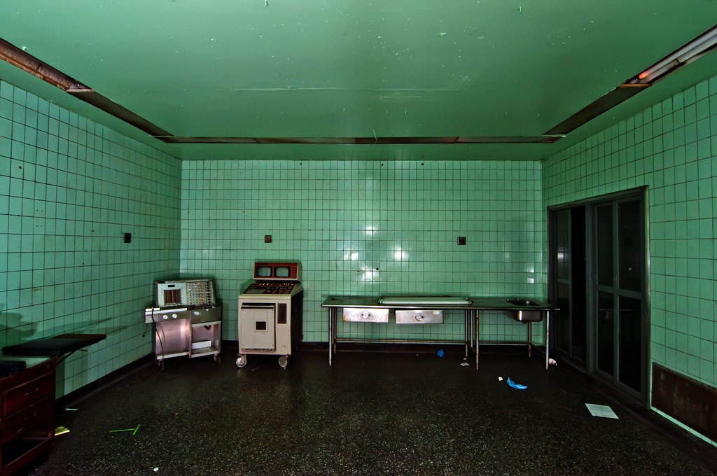 Photo shows a room with an operating table against an aqua wall. the hallway outside the room is dark

