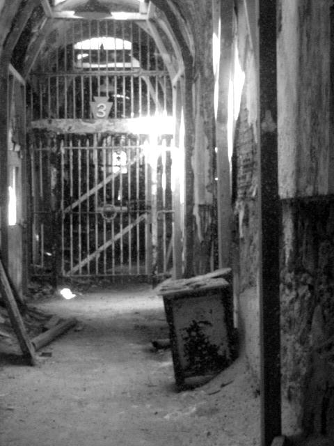 Eastern America's Most Haunted. Black and white photo of run down brick hallway with iron bars at the end with the number 3 above. 