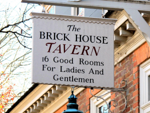 Off white wooden sign that reads The Brick House Tavern 16 Good Rooms For Ladies And Gentlemen
