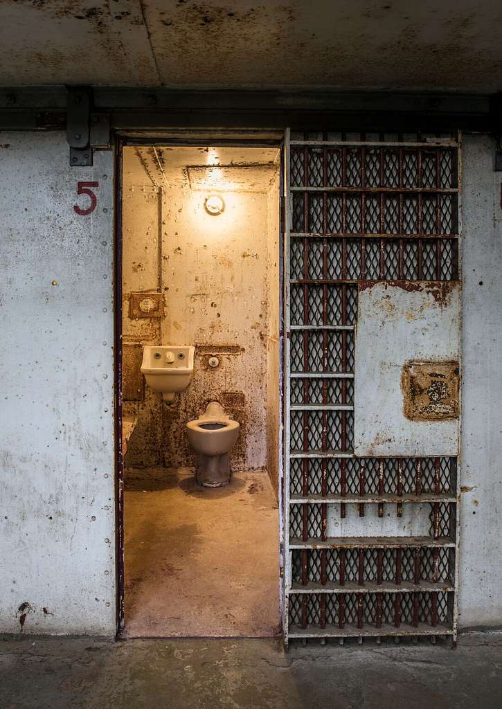 An iron prison cell. A light bulb hangs over a toilet and sink. The bars are open. The facade of the cell is very worn