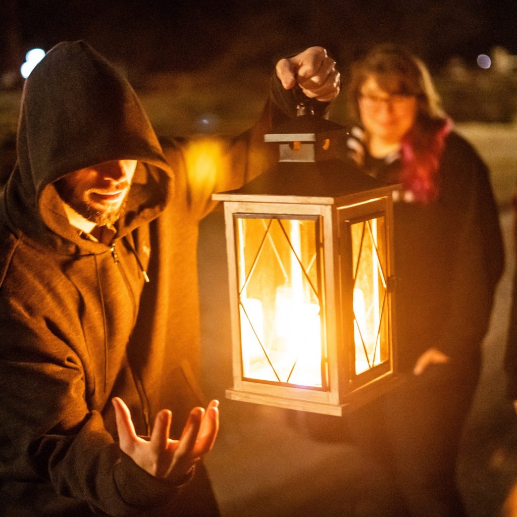A hooded tour guide sets the stage with his lantern as guests watch from the background at night