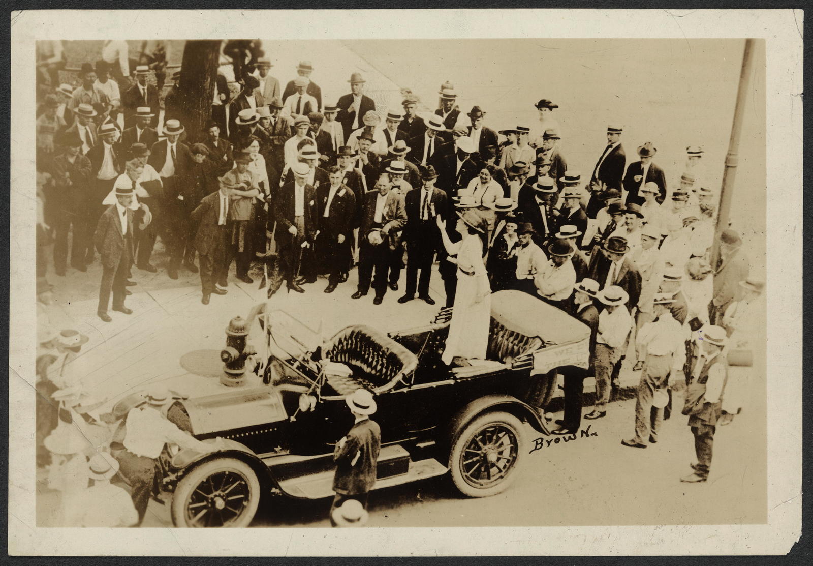Black and white photography of a many people gathered around a model T ford. Dressed in the 1920's style. 