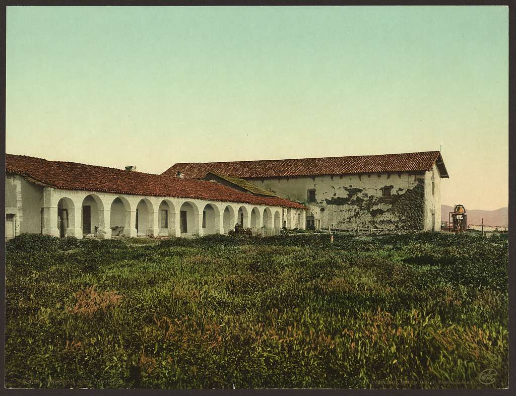 A spanish styled mission with red tile roofs and white walls sits in the brush during sunset