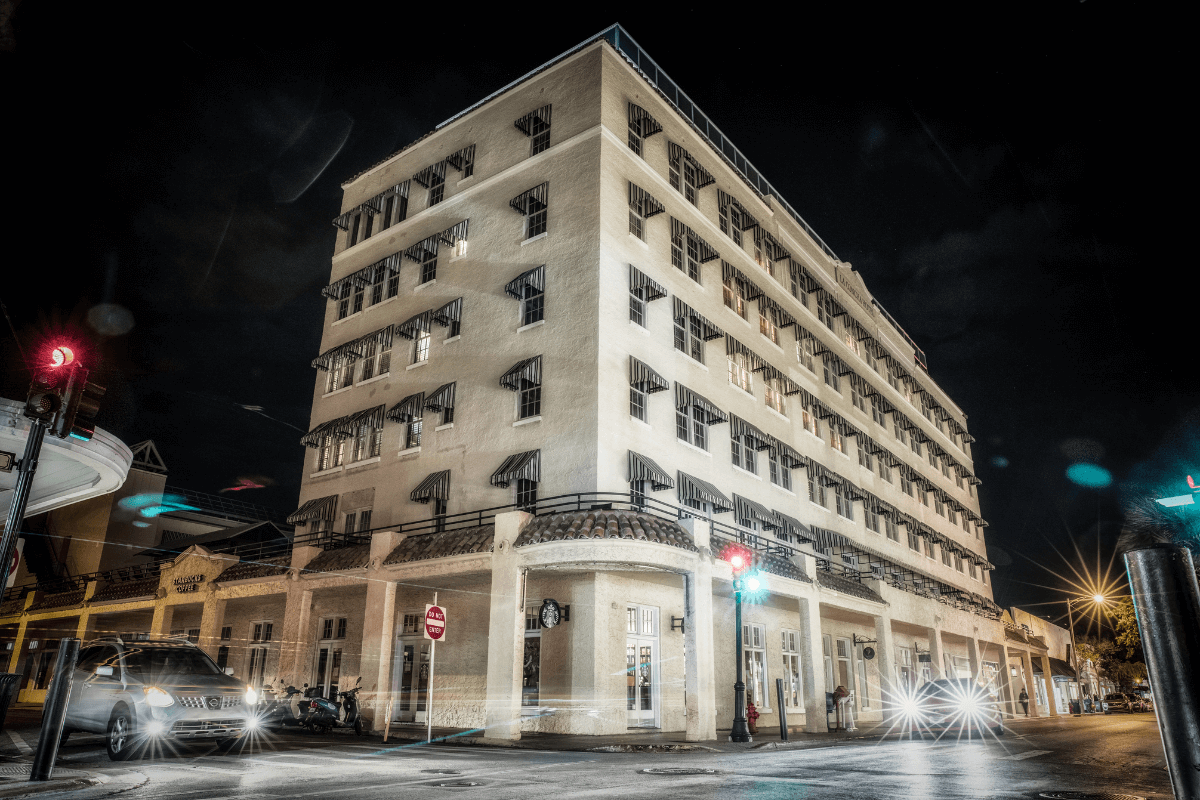 Multi level white brick hotel with black window awnings and metal fenced balcony at night.